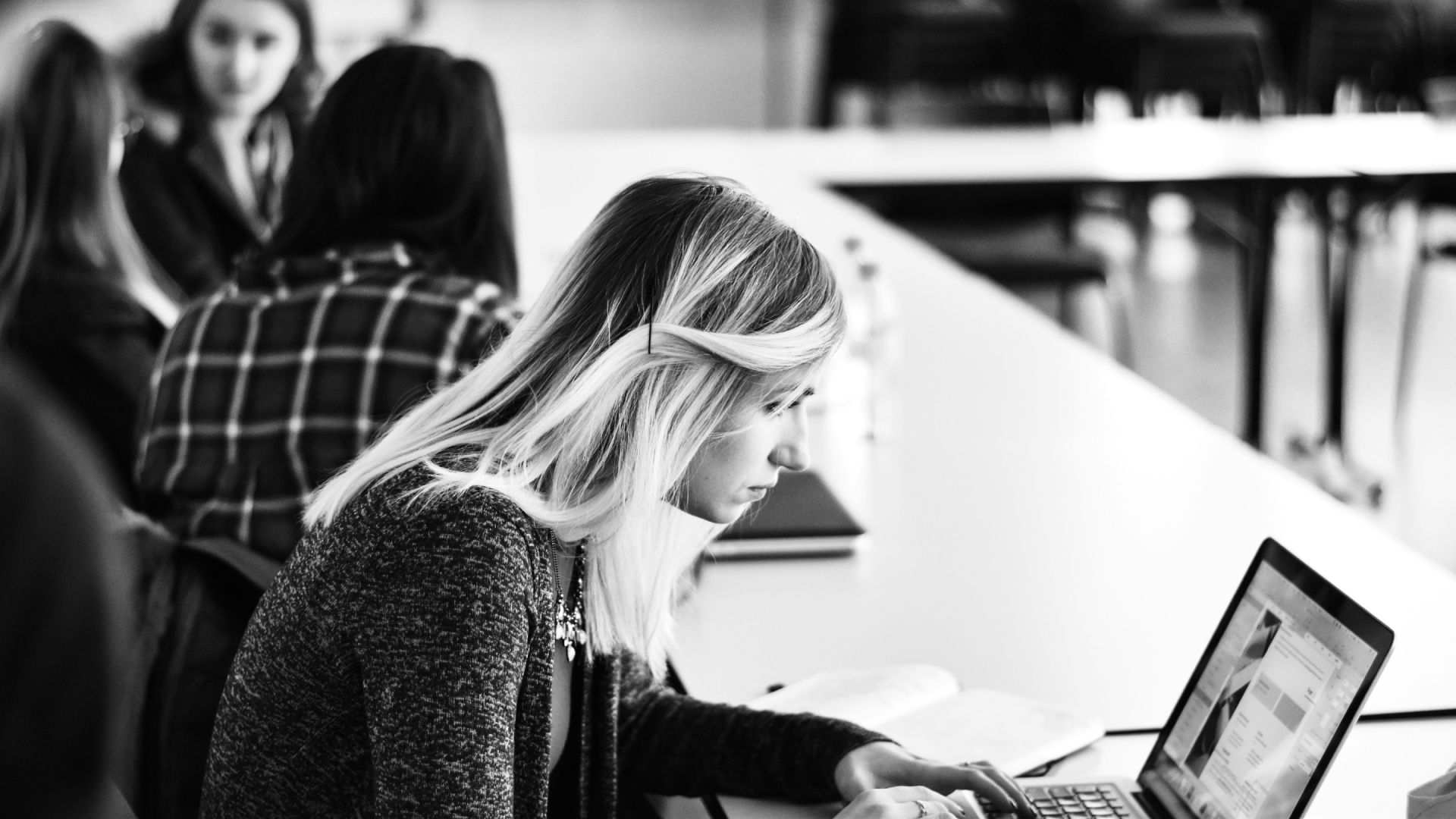 Woman working on laptop computer in school room with people in the background.