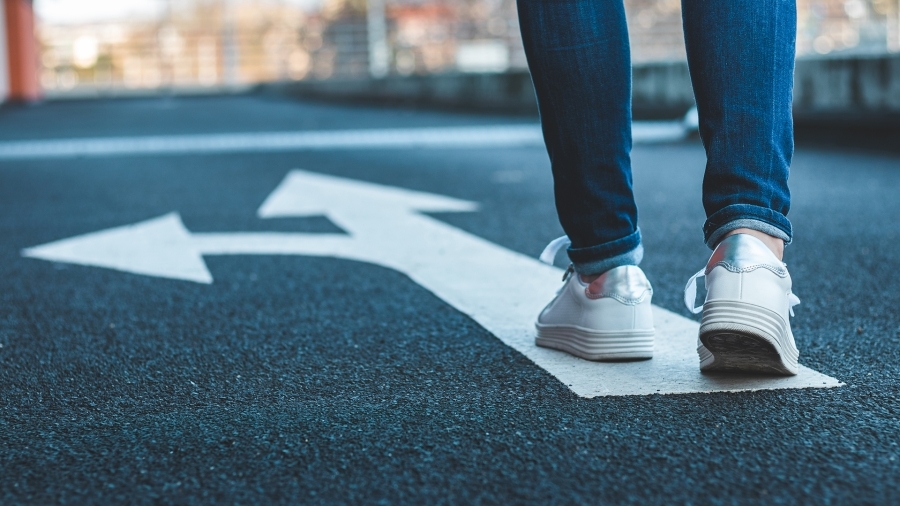 person's legs in jeans with white shoes walking on a road painted with arrows directed straight ahead and to the left