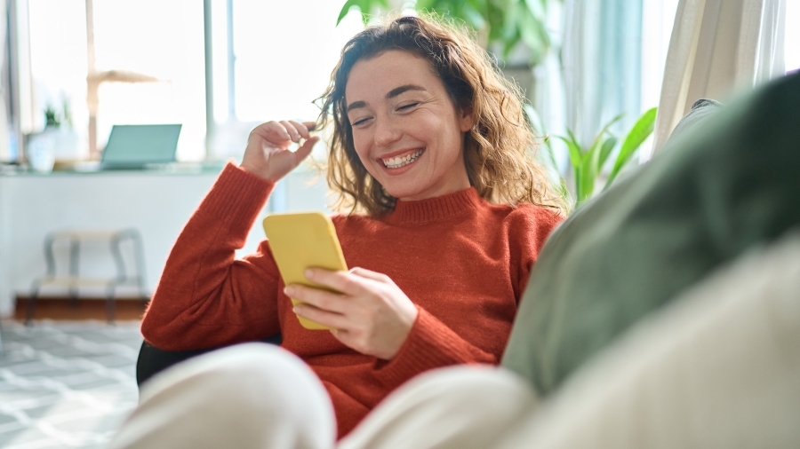 Une femme en pull rouge, souriant en regardant son téléphone.